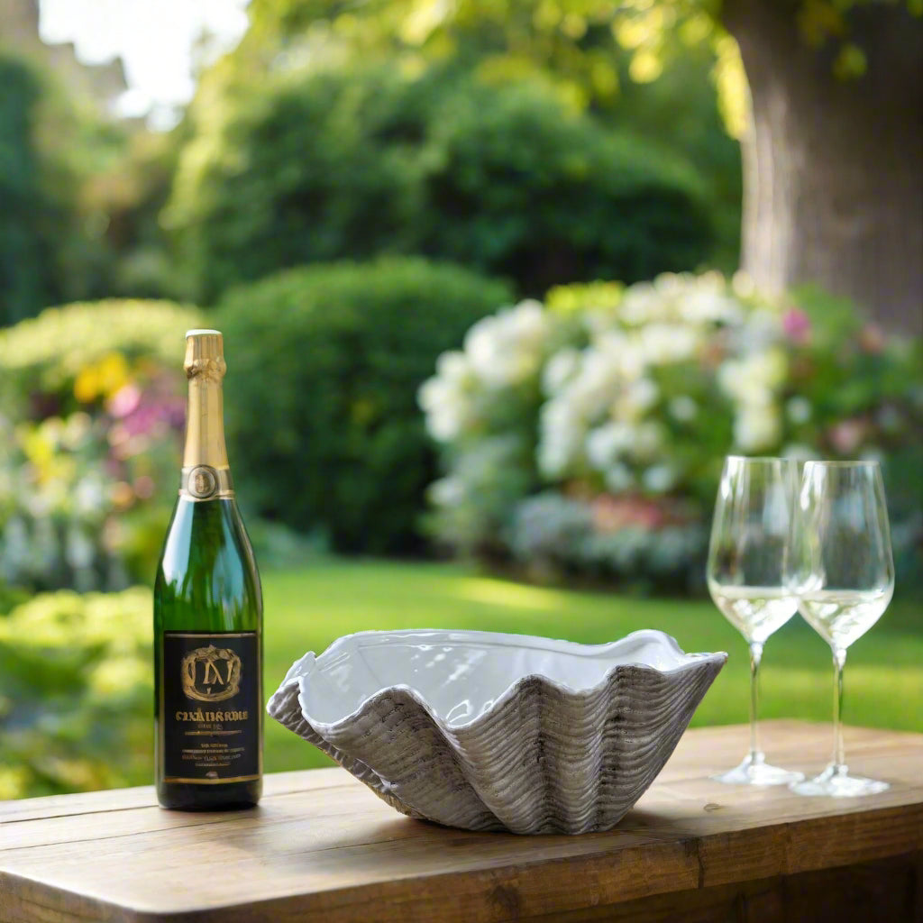 The large 40cm ceramic Adele Shell bowl used as a wine bucket, with a champagne bottle and two wine glasses visible on their side on a wooden outdoor table.