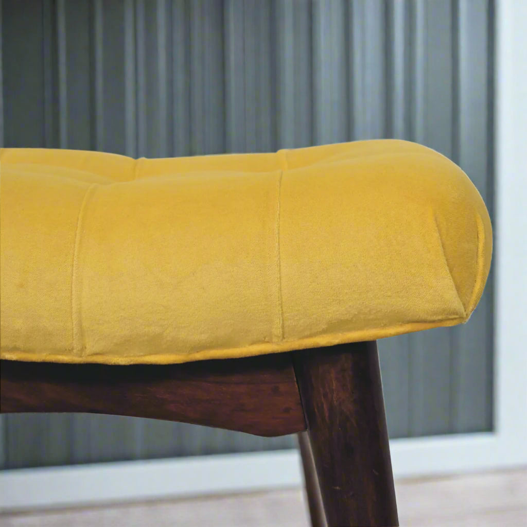 A closer look at the left edge of the mini mustard velvet curve stool, highlighting the upholstered seat and where it meets the wooden feet.