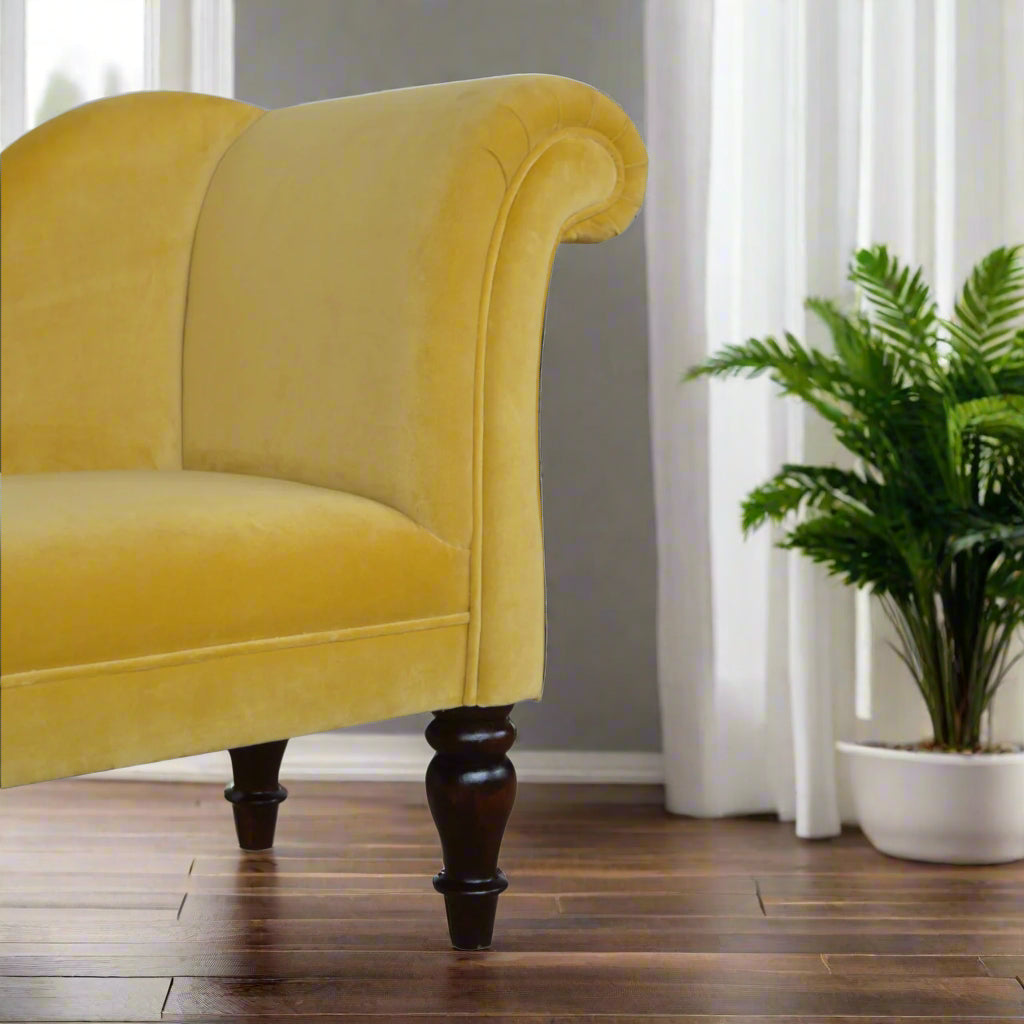 Almost eye-level view of the mustard velvet chaise's curved backrest, featuring an ornamental inhouse plant in the background and three visible turned legs.