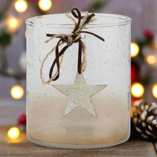 Front view of clear glass candle holder with a star-shaped ornament hanging from brown ribbon against a festive background with lights.