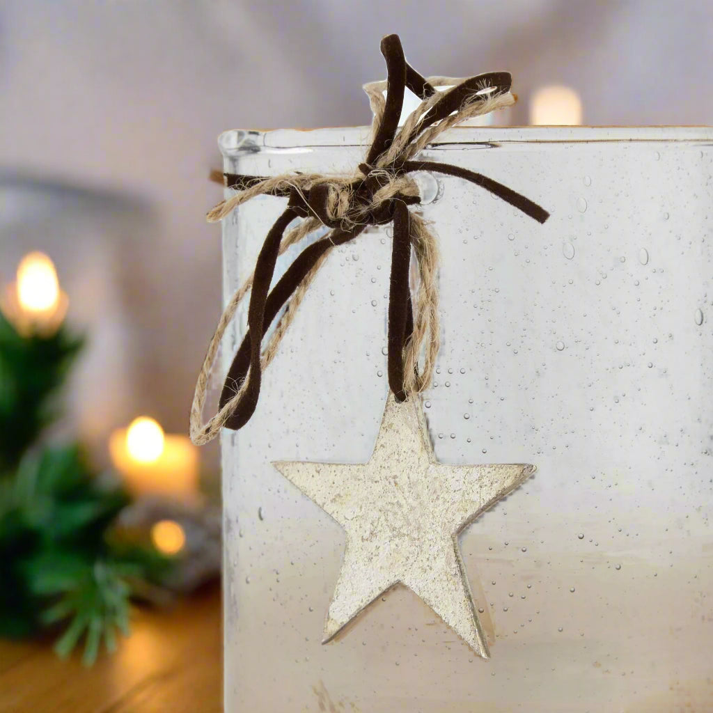 close view of clear glass candle holder with a star-shaped ornament hanging from brown ribbon against a festive background with lights.
