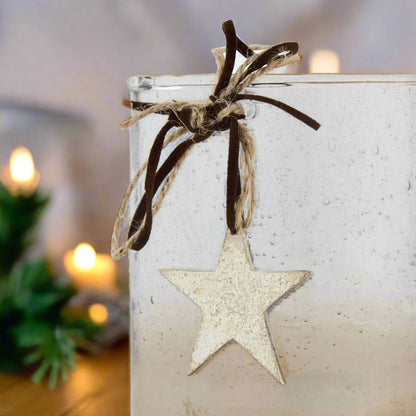 close view of clear glass candle holder with a star-shaped ornament hanging from brown ribbon against a festive background with lights.