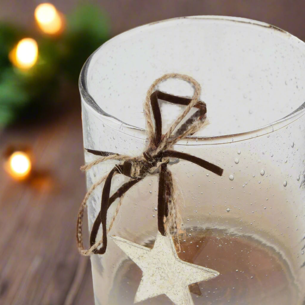 Clear glass candle holder with a star-shaped ornament hanging from brown ribbon against a festive background with lights.
