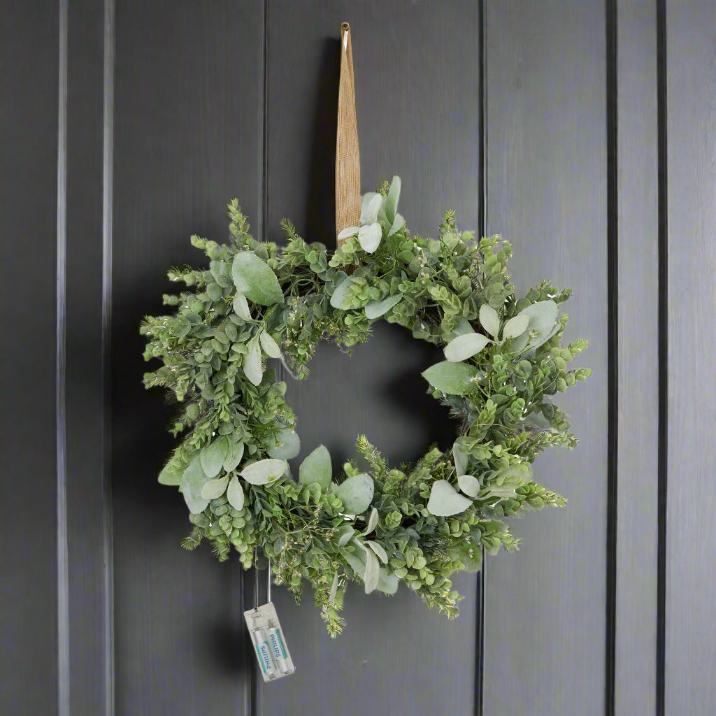 Stunning natural looking door wreath created with life-like Eucalyptus and Lambs ear greenery, hanging on a dark grey wood wall displaying the attached battery pack. Shop at Purple Flamingo UK