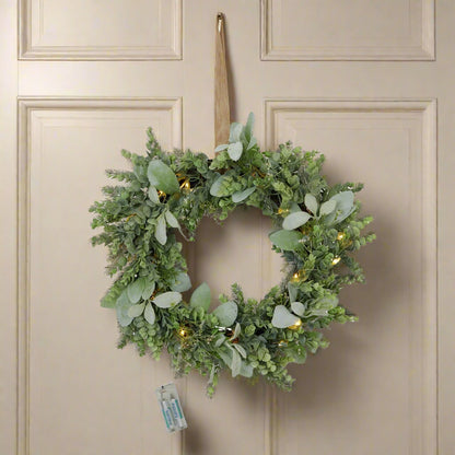 Stunning natural looking door wreath created with life-like Eucalyptus and Lambs ear greenery, the built-in LEDs are lit and glowing warmly, whilst the wreath is hanging on a cream wooden door, the attached battery pack is also displayed. Shop at Purple Flamingo UK!