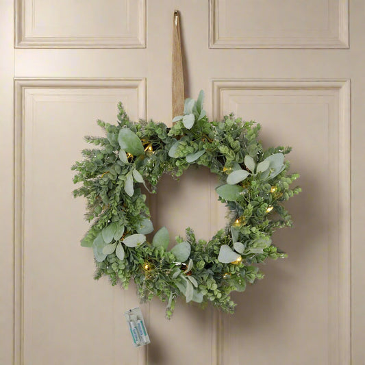 Stunning natural looking door wreath created with life-like Eucalyptus and Lambs ear greenery, the built-in LEDs are lit and glowing warmly, whilst the wreath is hanging on a cream wooden door, the attached battery pack is also displayed. Shop at Purple Flamingo UK!