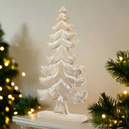 Decorative tiered wooden Christmas tree decoration with white wash snowy effect, placed on a cream mantle flanked by green garlands and Christmas tree bedecked with warm fairy lights. 