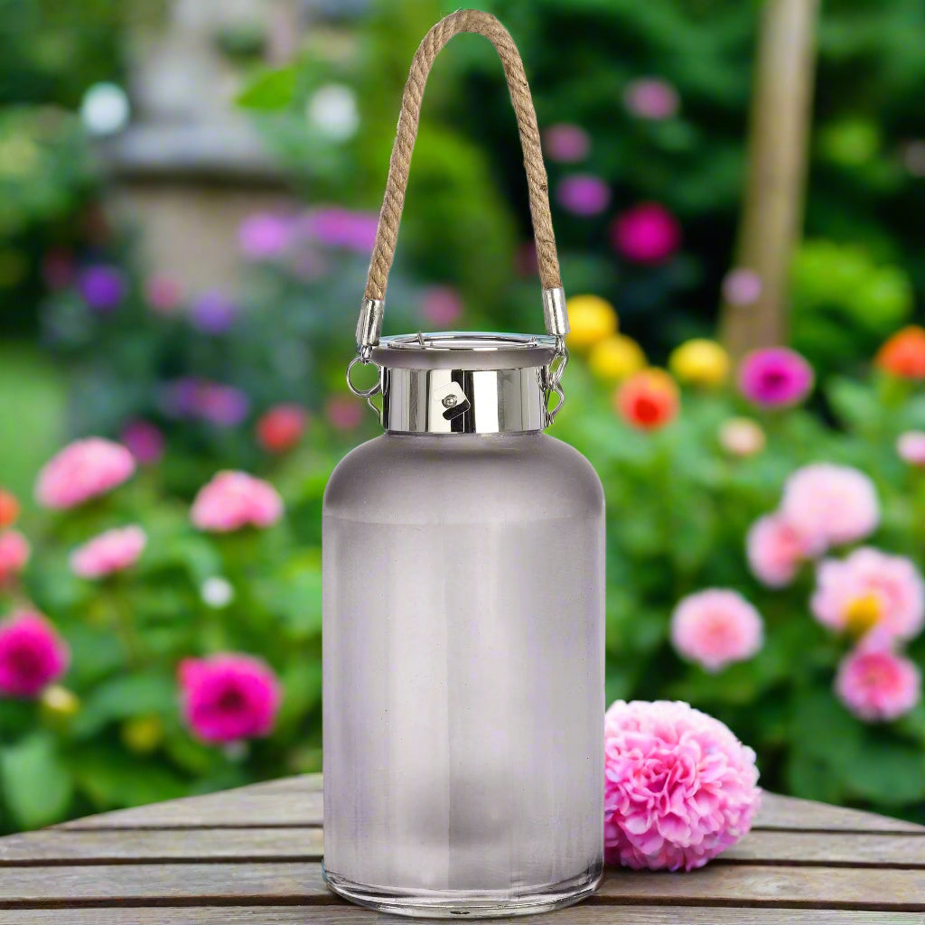 Lantern placed outdoors among flowers with visible rope handle for hanging.
