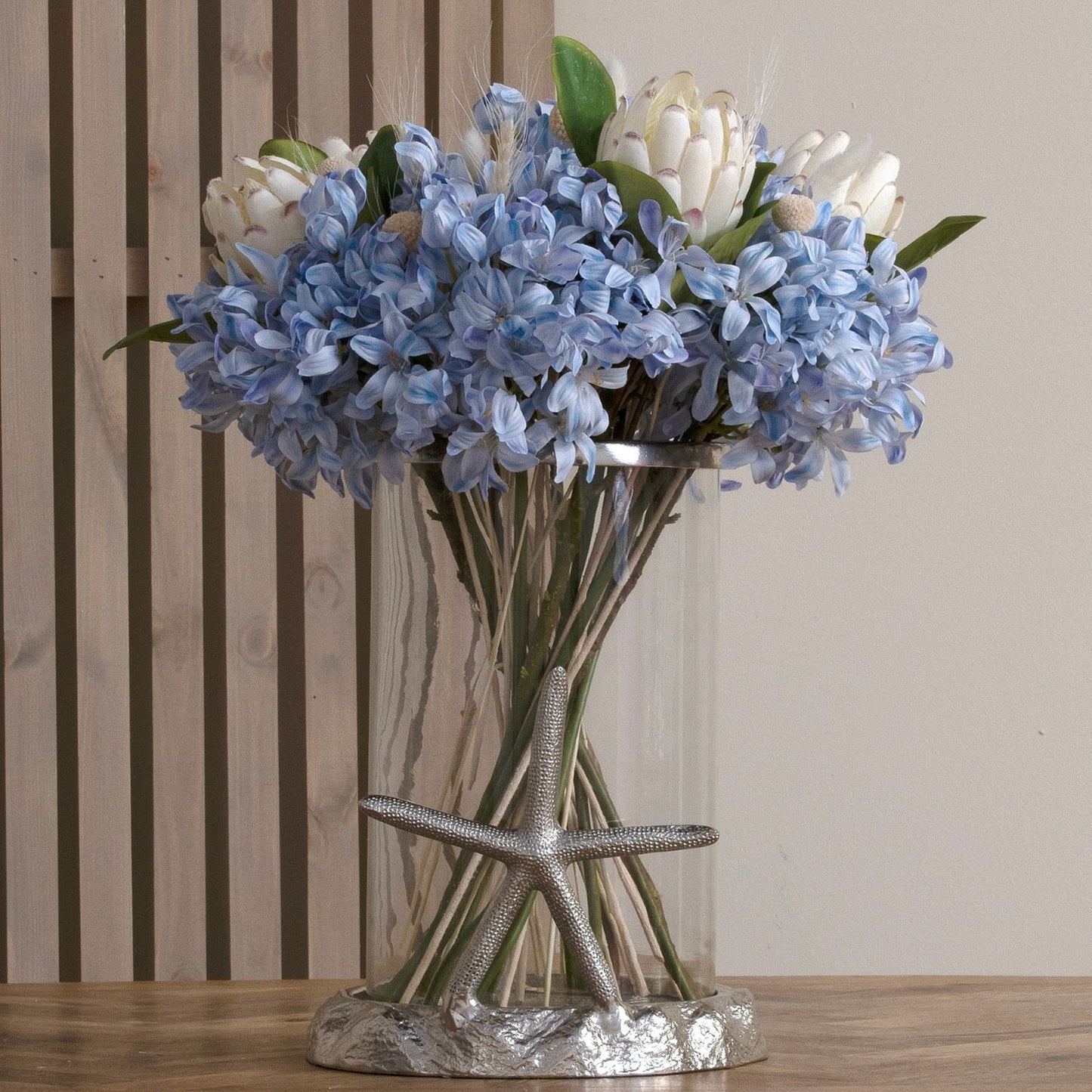 A bunch of light blue agapanthus in a clear vase beside a wooden wall.