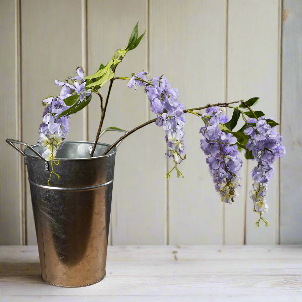 Two lilac wisteria stems in a stainless steel vase on a white surface.