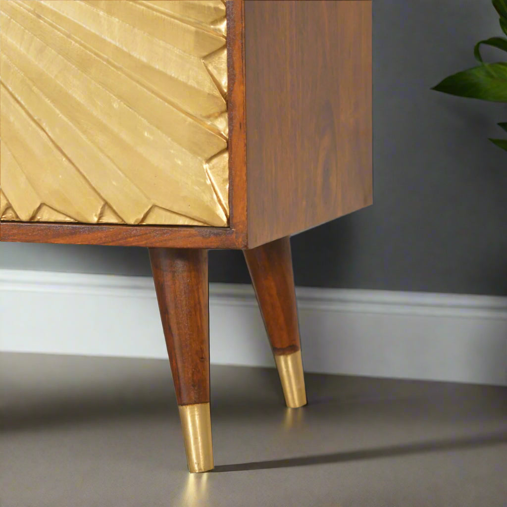 Close-up angled view of the Manila Gold Cupboard's tapered Nordic style legs with brass caps and a portion of its brass-plated sunburst door in a rich chestnut finish.