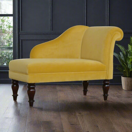Front angled view of the mustard velvet chaise lounge, showcasing its upholstered padded seat in yellow cotton velvet and the elegant turned legs, with an ornamental flower in the background.