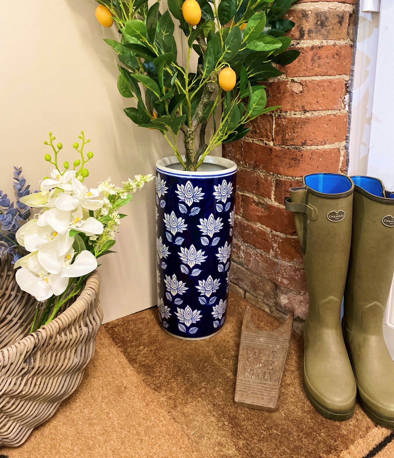 Purple Flamingo blue and white floral ceramic umbrella stand holding a lemon tree plant, styled in a hallway with a wicker basket and wellington boots.