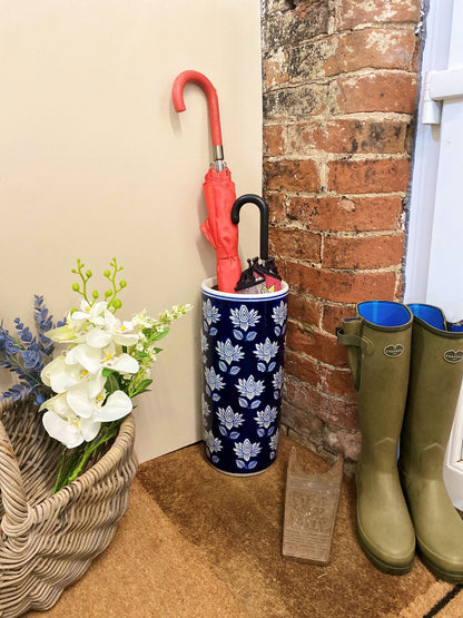 Purple Flamingo blue and white floral ceramic umbrella stand holding two umbrellas (red and black), positioned by a brick wall in a home entrance.