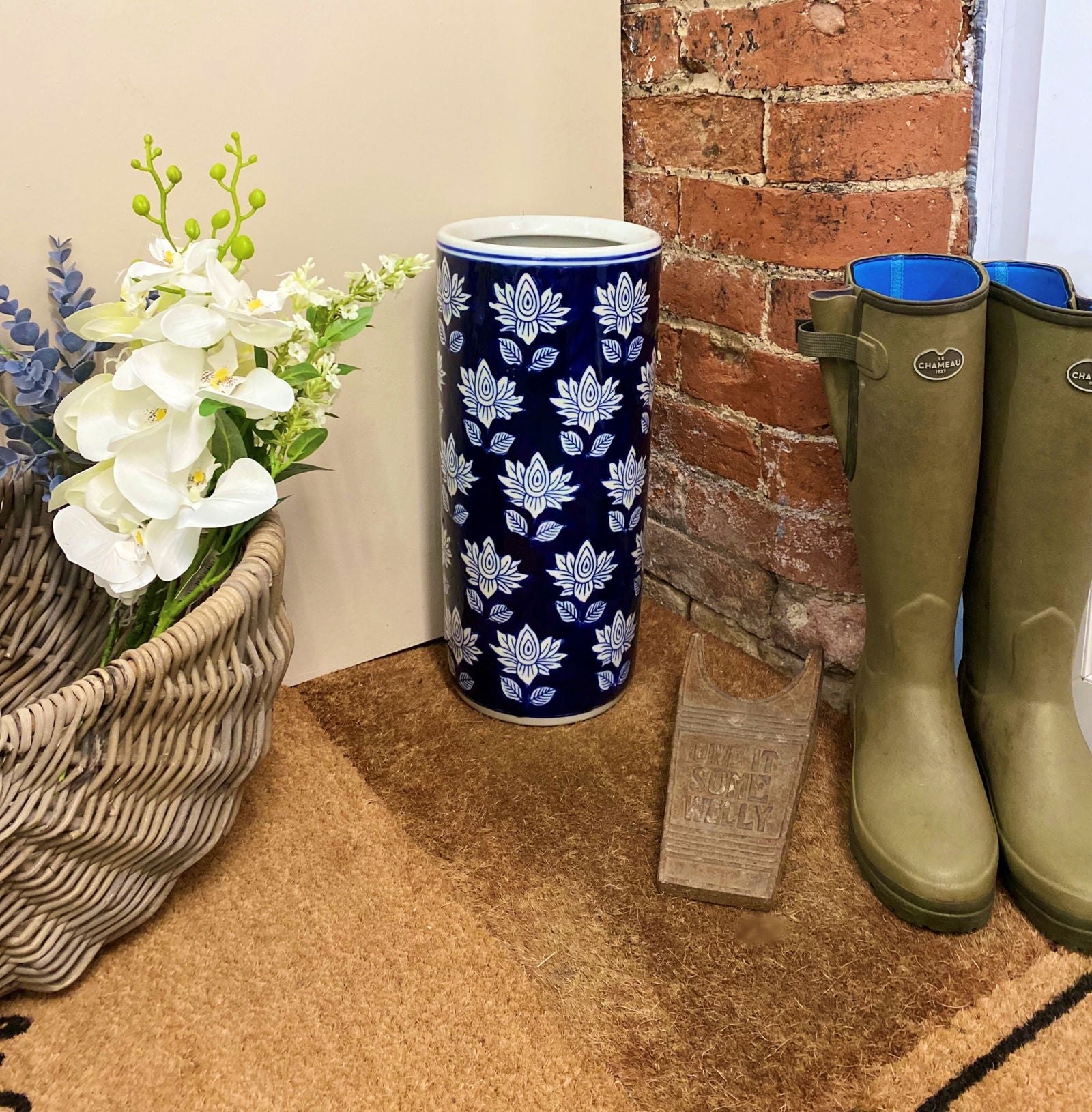 Purple Flamingo blue and white floral ceramic umbrella stand, styled in a hallway entrance with a wicker basket of flowers and green wellington boots.