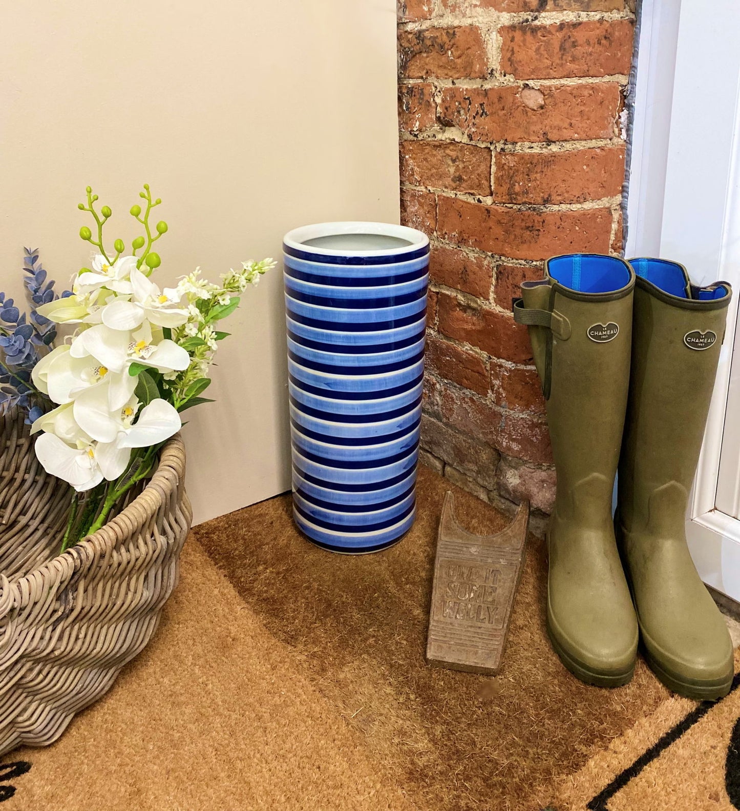 Purple Flamingo blue and white striped ceramic umbrella stand providing stylish storage in a hallway, next to a wicker basket and boots.
