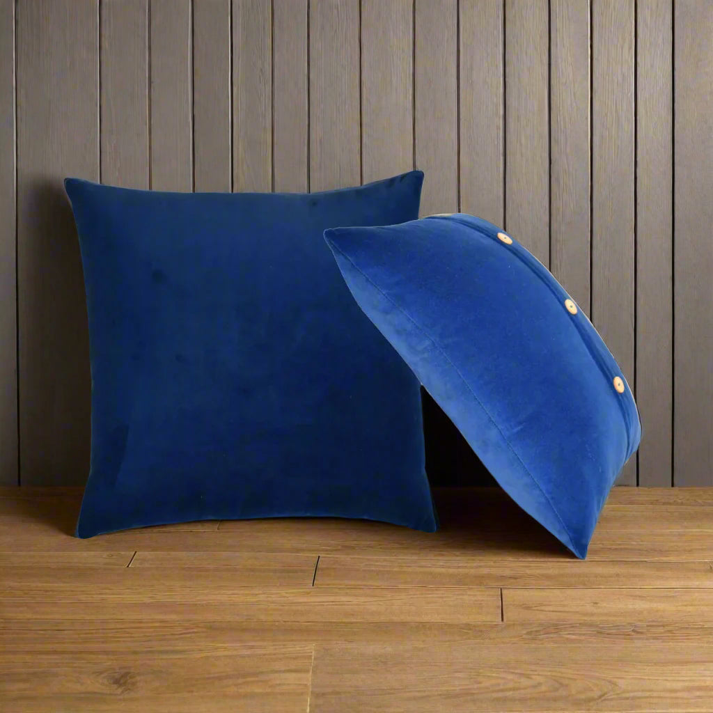 Two royal blue Quinn cushions in velvet, one leaning on the other, against a brown wall and wooden floor.
