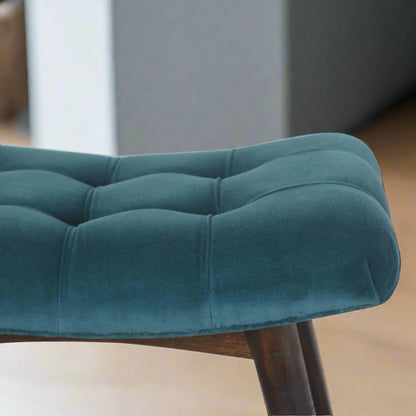 Elevated view of the left edge of the teal velvet curve stool, highlighting the luxurious velvet finish, deep button detailing, and the start of the wooden feet.