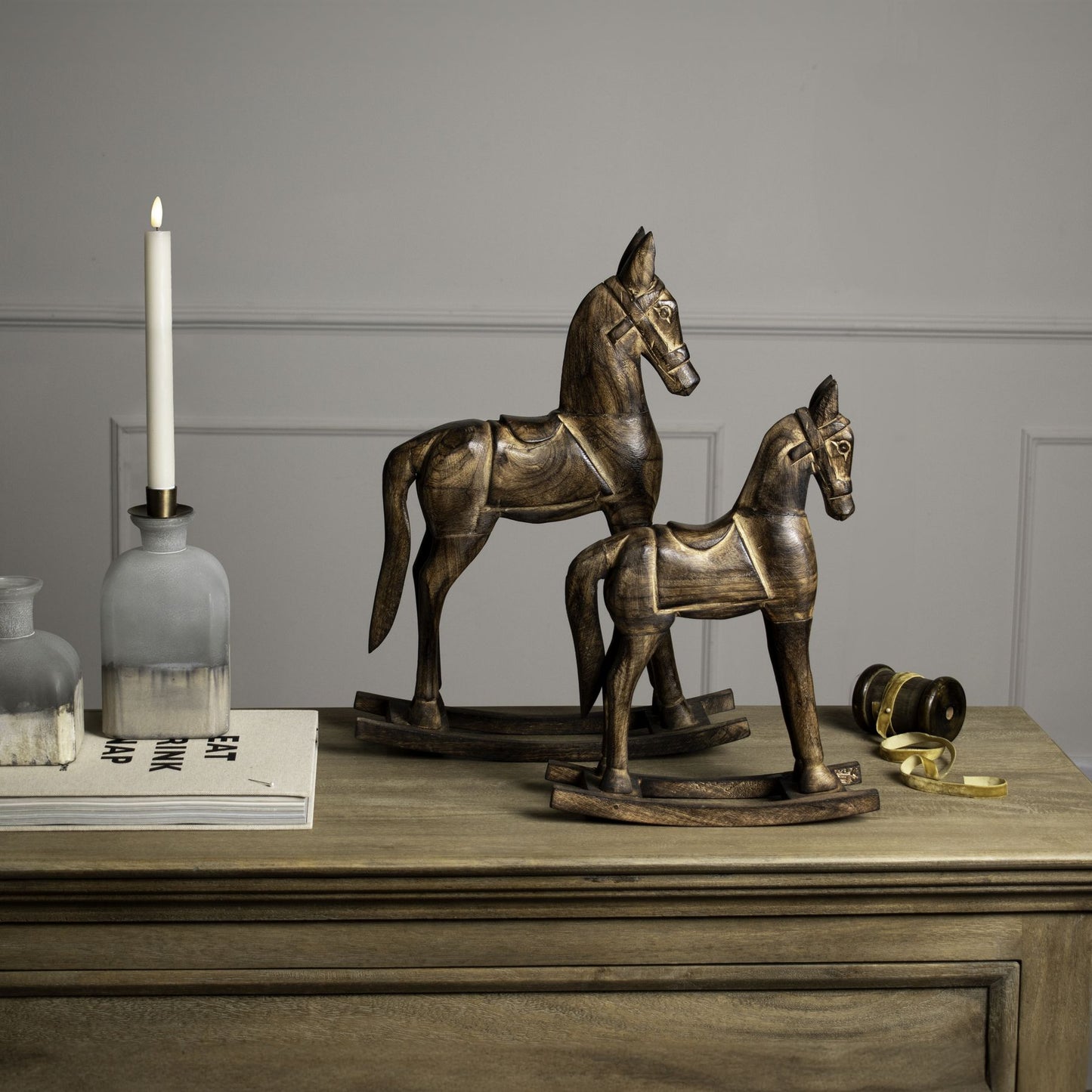 Two wooden rocking horse ornaments displayed side by side on a sideboard with a candle and book. 