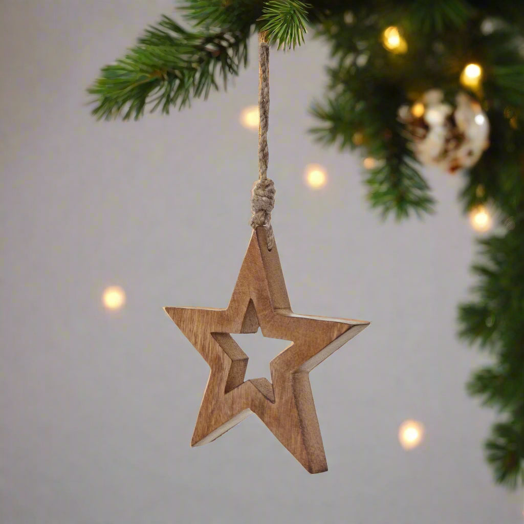 Wooden star shaped decoration with cut-out star middle hanging by the attached twine from a Christmas tree with fairy lights.